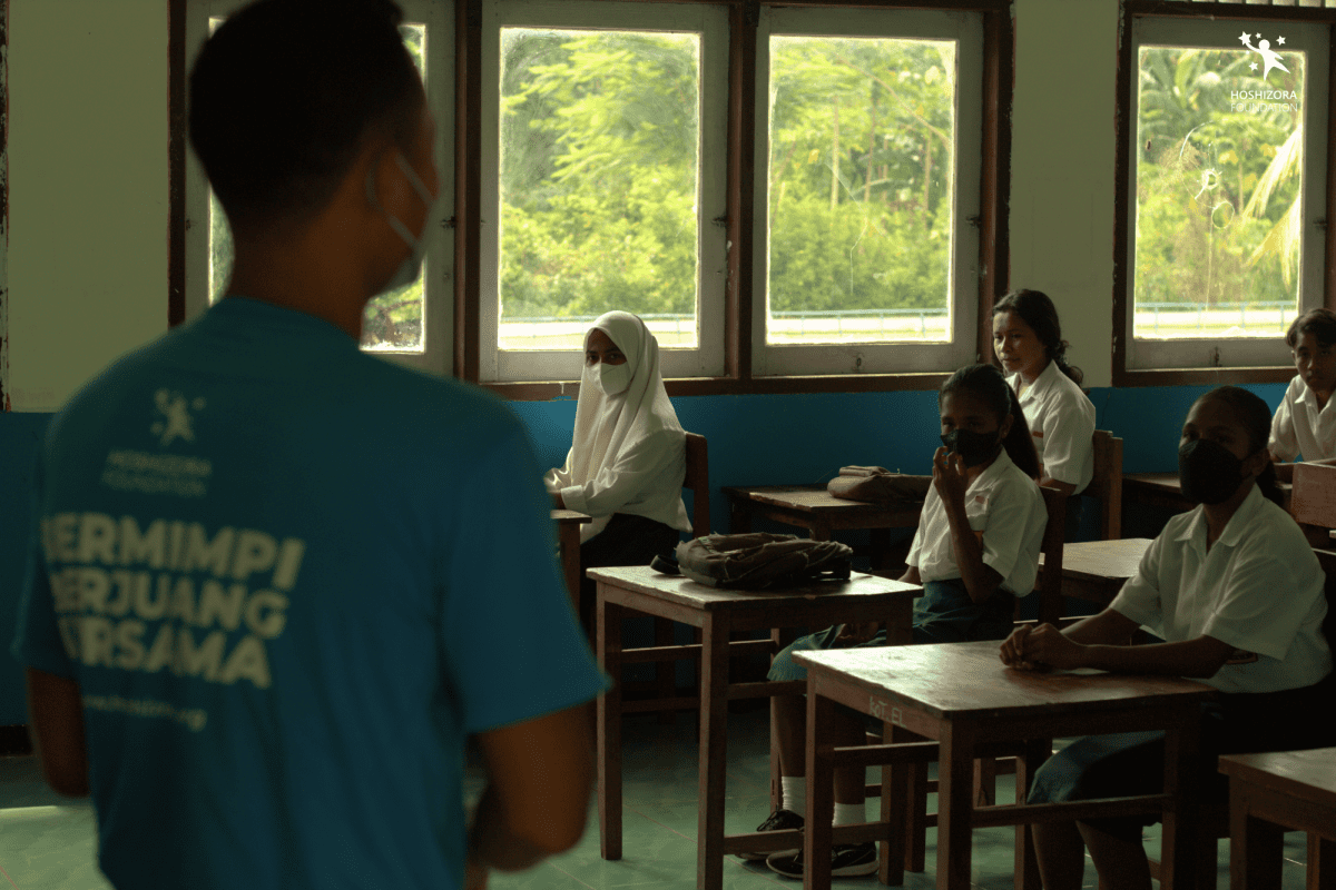 Foto menampilkan sebuah kelas sekolah dasar, dengan dinding putih yang bagian bawahnya berwarna biru. Terdapat empat jendela berukuran sedang di dinding yang terlihat. Di sebelah kiri bingkai (pada foreground), seorang staf Hoshizora berdiri menghadap murid-murid yang duduk di kursi meja masing-masing. Hanya tampak bagian belakang-samping dari si staf, yang memakai baju kaos biru bertuliskan BERMIMPI, BERJUANG, BERSAMA. Hari Guru Sedunia: Dampak Positif Donasi Pendidikan Hoshizora Foundation Terhadap Guru dan Anak Didik