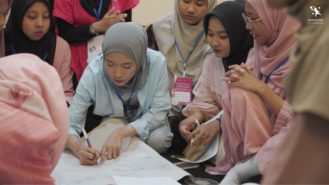 Adik-Adik Bintang Glow & Lovely sedang berkumpul untuk mengerjakan sebuah tugas di acara Capacity Building GAL 8. Seorang perempuan yang mengenakan kerudung abu-abu dan kemeja biru muda di tengah foto sedang menulis di sebuah kertas besar. Program Pengembangan Karakter di Hoshizora: Lebih dari Sekadar Beasiswa