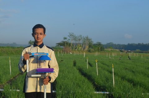 Adik ihsan sedang memegang alat Kapler X dengan latar belakang sawah